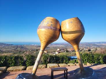Two giant wooden wine glass sculptures clink together with a scenic view of rolling hills, scattered houses, blue sky, and distant water on a bright, sunny day.