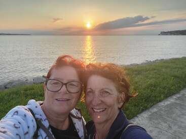 Two women smile for a selfie by a shoreline at sunset. Calm water, grassy bank, and a path are visible under a colorful, serene sky.