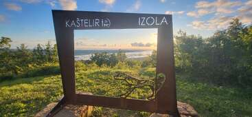 A metal frame labeled "Kaštelir Izola" overlooks a scenic landscape with lush greenery, sea, and sky. A dinosaur figure is cut out below.