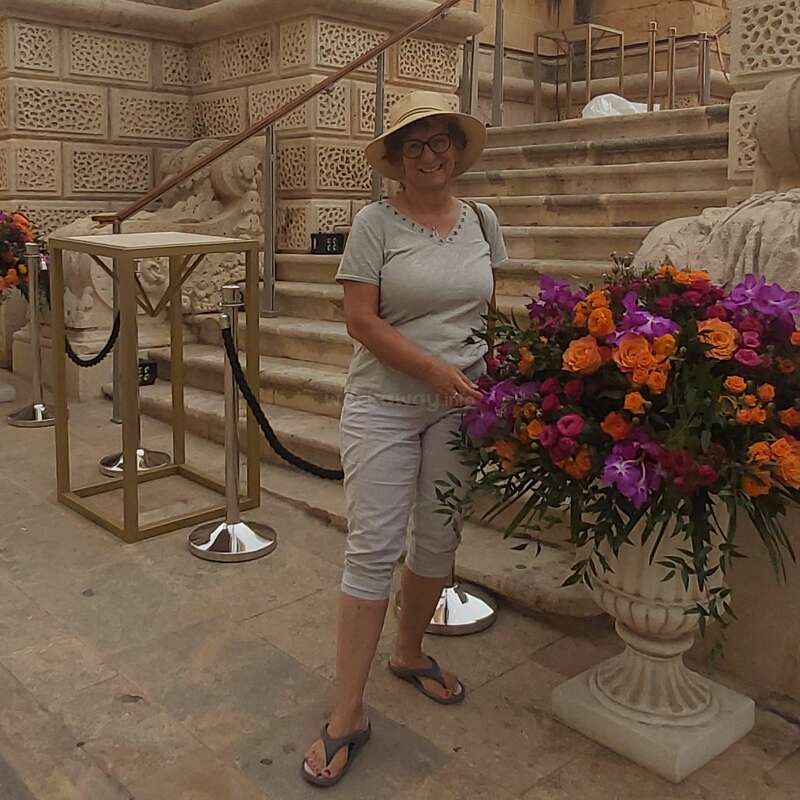 Eine Frau mit Hut und Brille steht lächelnd neben einem großen, bunten Blumengesteck. Sie steht vor einer Steintreppe mit verzierten Mauern dahinter.