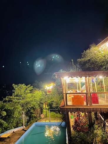 Das Bild zeigt eine ruhige nächtliche Szene mit einem Pool und einer überdachten Holzterrasse, umgeben von üppigem Grün und Bäumen, vor einem dunklen Himmel.