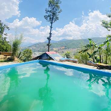 La imagen representa una serena escena con una piscina de un azul intenso, rodeada de exuberante vegetación y una cordillera lejana, sobre un cielo azul brillante con nubes blancas.