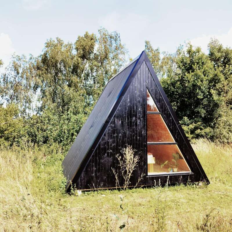 L'image représente une petite cabane noire à cadre en A avec une fenêtre triangulaire, située dans un champ herbeux entouré d'arbres et d'un ciel bleu avec des nuages blancs.