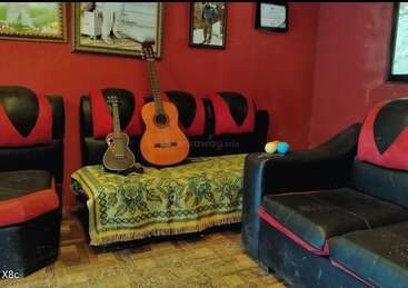 A cozy living room with red walls, black and red sofas, guitars resting on a covered ottoman, framed pictures on the wall, and colorful juggling balls.