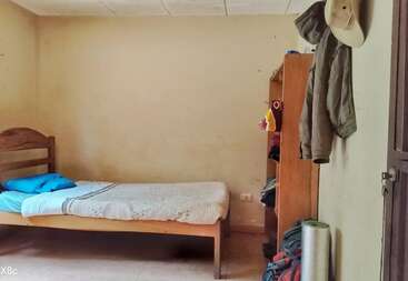 A simple room with a single wooden bed, blue pillows, blanket, shelf with clothes, jackets hanging on the wall, hat, and plain beige walls.