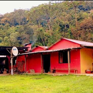 This image shows a rustic red and yellow house with a satellite dish, surrounded by lush green forest and hills, creating a peaceful rural atmosphere.