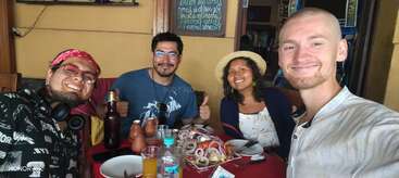 Four friends sit at a restaurant table, smiling at the camera. Drinks and seafood are on the table. The atmosphere is cheerful and relaxed, with casual attire.