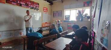 A teacher stands at the whiteboard, instructing attentive children seated at desks in a classroom decorated with colorful posters, educational charts, and a sunny window.