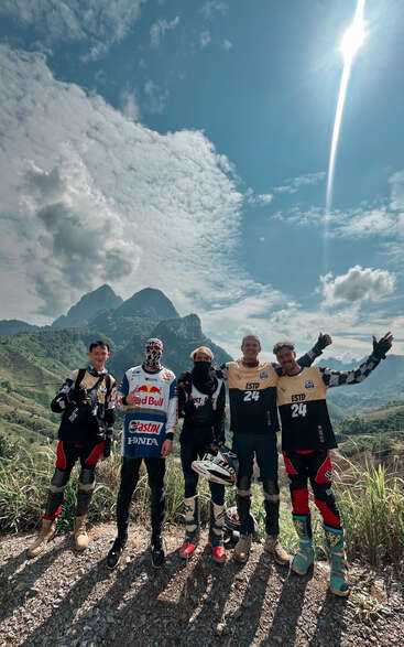Cinco pilotos de motocross posam em uma trilha de montanha panorâmica sob um céu ensolarado, usando equipamentos, sorrindo, com colinas verdejantes e picos dramáticos ao fundo.