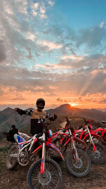 Uma pessoa com equipamento de motocross fica ao lado de motos de terra ao pôr do sol, com montanhas e um céu dramático, criando uma sensação de aventura e liberdade na natureza.