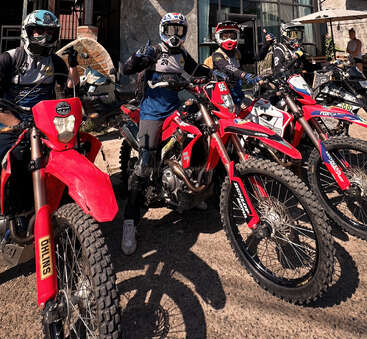 Quatro pilotos de motocross com equipamentos e capacetes posam em motos de terra vermelhas em frente ao Rabica Café. A atmosfera é aventureira, ensolarada e energética, com arquitetura moderna.