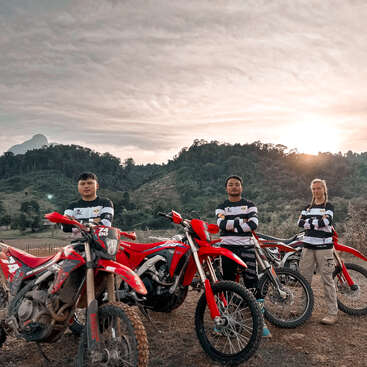 Three people stand confidently beside their red dirt bikes on a hilltop, wearing matching shirts, with mountains and a dramatic sunset sky in the background.