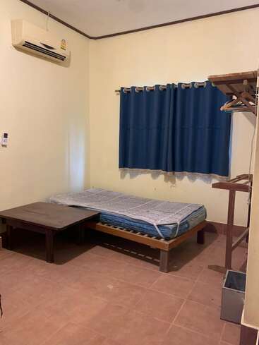 Esta imagem mostra um quarto simples e pouco mobiliado com uma cama de solteiro, cortinas azuis, ar-condicionado, mesa de madeira, cabideiro e piso de azulejos. As paredes são lisas.