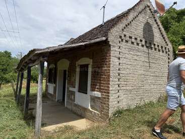 Uma casa velha e rústica de tijolos com o telhado danificado fica em meio a uma vegetação alta. Um cara de bermuda e chapéu passa pela frente, parcialmente visível.