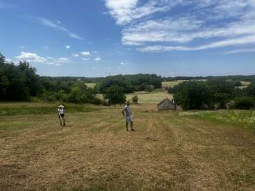 Duas pessoas estão em um campo aberto e ensolarado, cercado por árvores verdes e casas distantes, sob um céu azul com nuvens brancas e finas. Uma paisagem tranquila.