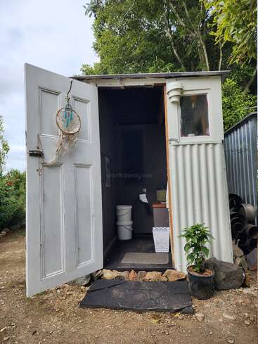 Das Bild zeigt ein kleines, weißes Gebäude mit einer offenen Tür, in dem sich eine Toilette, ein Waschbecken und eine Topfpflanze befinden, in einem Hof oder Garten.