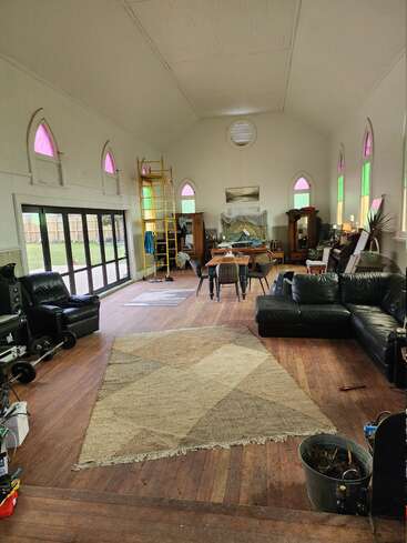 Dieses Bild zeigt einen geräumigen Raum mit hohen Decken, Buntglasfenstern, verstreuten Möbeln wie Sofas und Tischen, einem Holzboden und einer Leiter im hinteren Bereich.