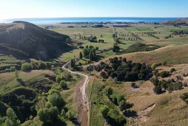 Des collines couvertes d'herbe verte et d'arbres entourent un chemin de terre sinueux. Au loin, des terres agricoles plates s'étendent vers l'horizon bleu étincelant de l'océan.