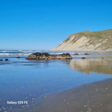 Eine ruhige Küstenszene mit klarem, blauem Himmel, sanften Wellen, reflektierendem, nassem Sand, felsigen Ausläufern und einer großen Klippe. In der unteren Ecke steht "Galaxy S20 FE".