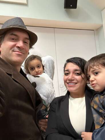 Eine glückliche vierköpfige Familie posiert in einem Haus. Der Vater trägt einen Hut und einen braunen Mantel, die Mutter lächelt, die Tochter hat Engelsflügel, und der Sohn schaut neugierig.