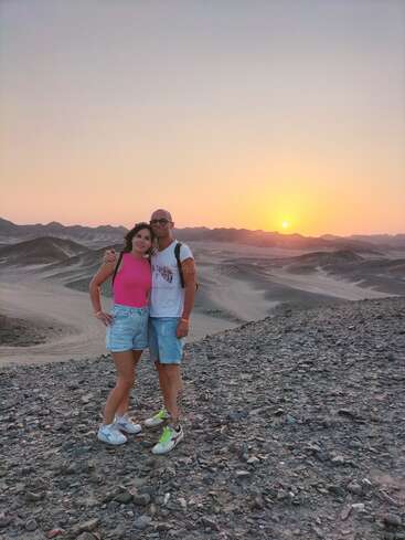 Una pareja se encuentra en un terreno rocoso desértico al atardecer, con colinas ondulantes al fondo. Sonríen, visten ropa informal de verano y disfrutan del hermoso paisaje.