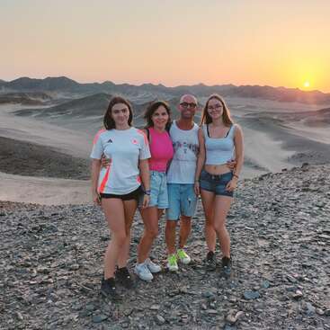 Quatre personnes posent ensemble sur un terrain désertique et rocailleux au coucher du soleil, avec des montagnes en arrière-plan. Ils portent des vêtements d'été décontractés et profitent du paysage naturel à couper le souffle.