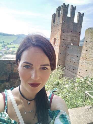 Eine Frau macht ein Selfie vor einer alten Steinburg mit hohen Türmen. Im Hintergrund sieht man grüne Felder, Hügel und einen hellen, klaren Himmel.