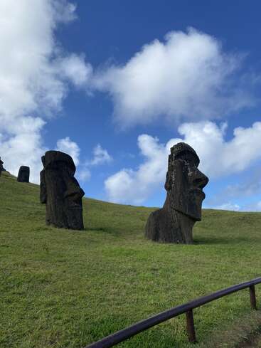 Deux statues Moai en pierre massive se dressent sur une colline herbeuse sous un ciel d'un bleu éclatant, parsemé de nuages blancs. Une rambarde métallique longe le premier plan.