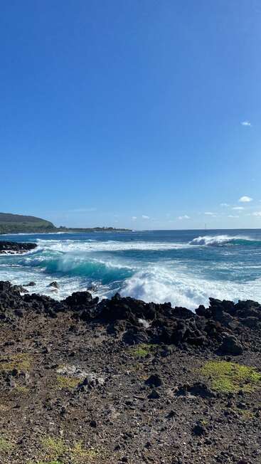 Les vagues s'écrasent sur les roches volcaniques noires sous un ciel d'un bleu éclatant. Des collines lointaines, des taches vertes et de légers nuages créent un paysage côtier serein et d'une grande beauté naturelle.