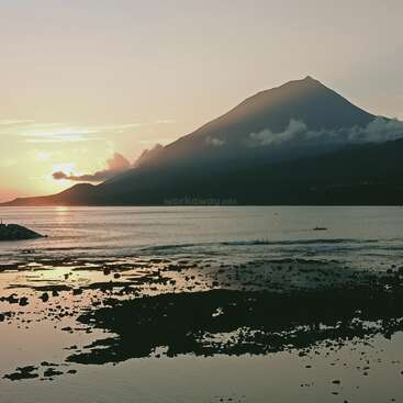 Um pôr do sol tranquilo atrás de uma grande montanha coberta de nuvens reflete lindamente nas águas calmas. A costa rochosa em primeiro plano realça a atmosfera serena e pacífica da natureza.