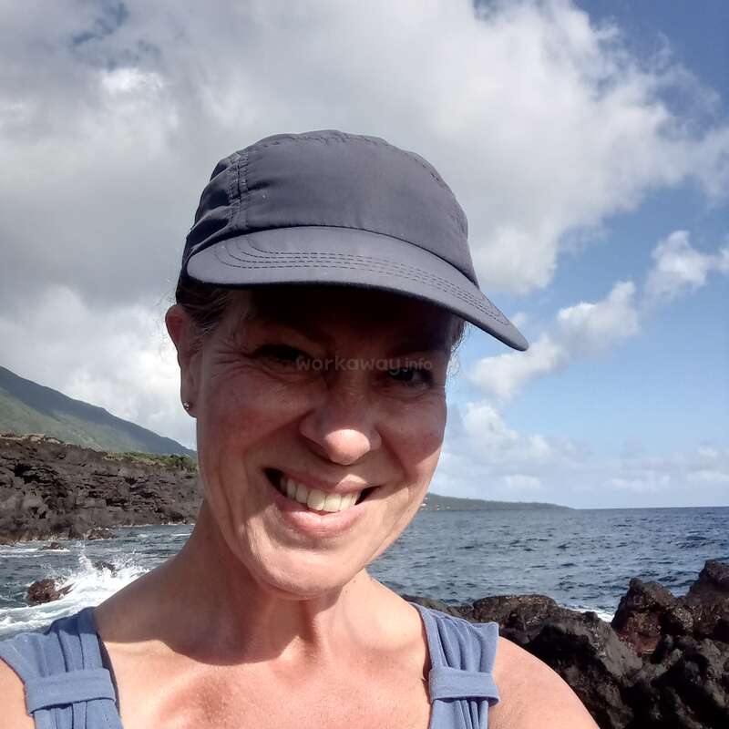 Uma mulher usando um boné cinza sorri em uma selfie tirada à beira-mar, junto às rochas. O céu está parcialmente nublado, e as ondas quebram nas rochas atrás dela.