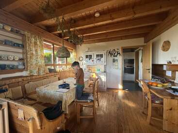 Una acogedora cocina de madera, bañada por la luz del sol y con vigas a la vista, un niño sentado a la mesa, estantes abiertos con tazas, hierbas aromáticas colgadas, fruta y una decoración cálida y hogareña por todas partes.