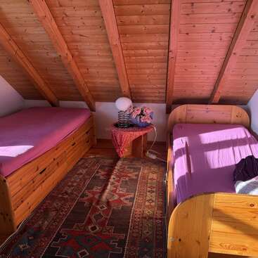 Esta acogedora habitación en el ático cuenta con dos camas de madera con sábanas rosas, una alfombra con motivos coloridos, un techo inclinado de madera, una mesita con una lámpara y un arreglo floral.