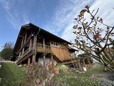 Una casa rústica de madera con una terraza elevada se alza sobre una ladera cubierta de hierba. En primer plano, bajo un cielo parcialmente nublado, se ven plantas con flores y un árbol sin hojas.
