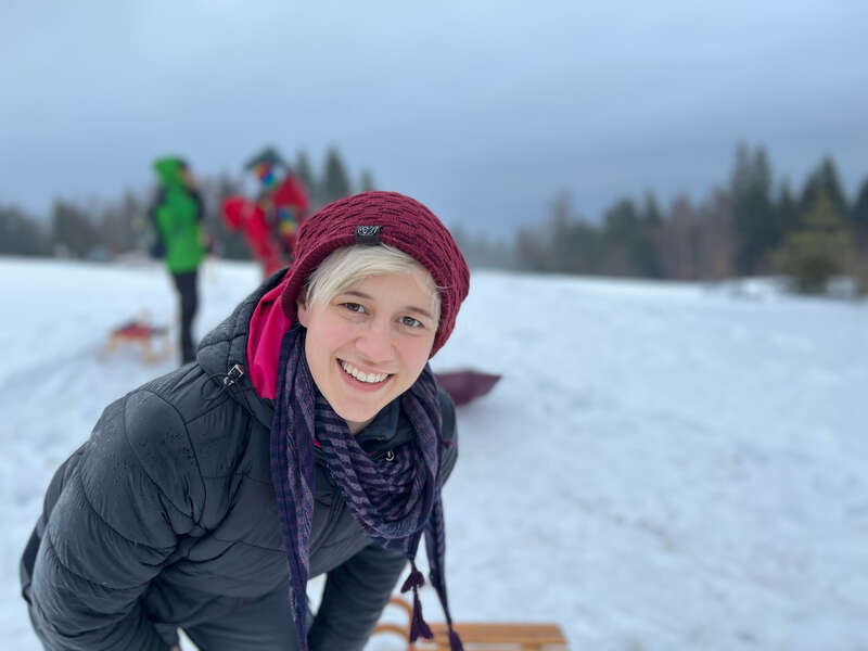Una persona sonriente, con un gorro granate y una chaqueta de invierno, disfruta del aire libre nevado, con figuras borrosas y trineos al fondo. El ambiente parece divertido y animado.