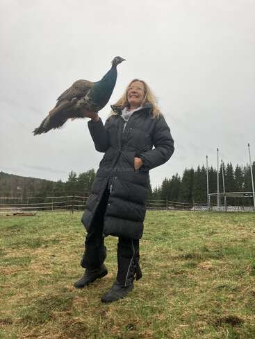 Eine lächelnde Frau in einem langen schwarzen Mantel steht draußen auf dem Rasen und hält einen Pfau auf dem Arm. Im Hintergrund sind Bäume und ein bewölkter Himmel zu sehen.