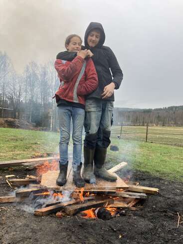 Zwei Kinder stehen nebeneinander auf Holzbrettern über einem Lagerfeuer, sie tragen Jacken und Stiefel und sind von Rauch umgeben, im Hintergrund sieht man eine grasbewachsene, ländliche Landschaft.