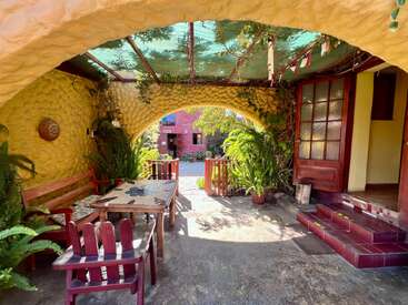 Eine gemütliche, sonnendurchflutete Terrasse mit rustikalen Holzmöbeln, Topfpflanzen, gewölbten gelben Wänden und einem grünen Baldachin, die eine einladende, ruhige und gemütliche Atmosphäre im Freien schafft.