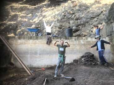 The image depicts four individuals standing in front of a concrete wall, with two people on top and two below, amidst a rocky terrain with tools scattered around.