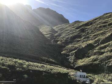 The image depicts a serene mountainous landscape with a white building at its base, set against a clear blue sky with the sun shining brightly in the top left corner.
