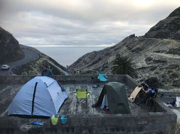 The image depicts a serene camping scene on a concrete platform, surrounded by mountains, with a tent, chairs, and bicycles, set against a cloudy sky.
