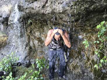 The image depicts a man standing in front of a rocky cliff, with water cascading down behind him, as he holds his head under the flow.