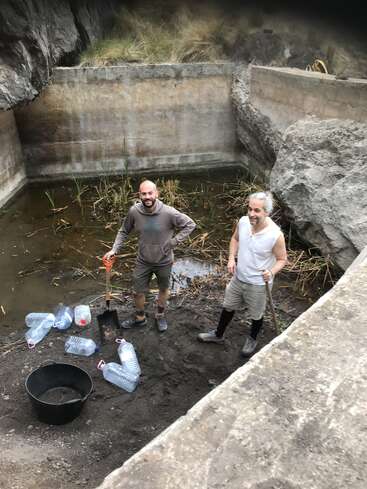 Two men stand in a muddy area, surrounded by water bottles and a bucket, with shovels in hand, near a concrete wall and rocks, in a natural setting.