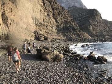 The image depicts a group of people walking along a rocky beach, with a large cliff looming in the background and a body of water to the right.