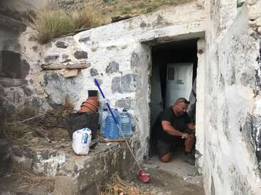 A man is kneeling in a doorway of a stone building, with cleaning supplies and water bottles on a ledge to his left. He is working on something.