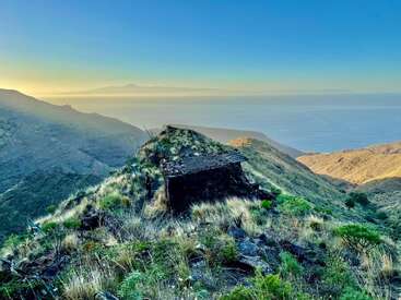 The image depicts a rugged, grassy hillside with a small, dilapidated structure, overlooking a vast body of water and distant mountains under a clear blue sky.