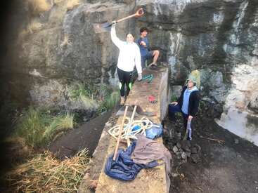 The image depicts four individuals standing on a stone bridge, surrounded by tools and clothing, with a rugged cliff face serving as the backdrop.