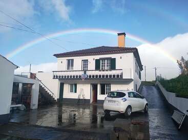 The image depicts a two-story white house with a brown roof, a white car parked in front, and a vibrant rainbow arcing over the top of the building.