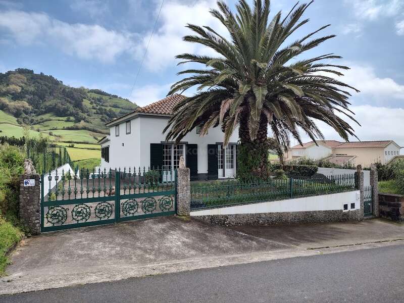 The image depicts a white house with a tiled roof, a palm tree, and a green fence, set against a backdrop of rolling hills and a blue sky.