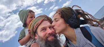 Um momento feliz em família: um cara de barba sorri, uma mulher dá um beijo na bochecha dele e uma criança pequena anda nos ombros dele contra um céu ensolarado.